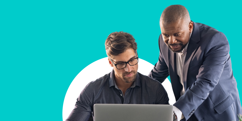A man, leaning over and helping another man as he sits at a desk, working on his laptop