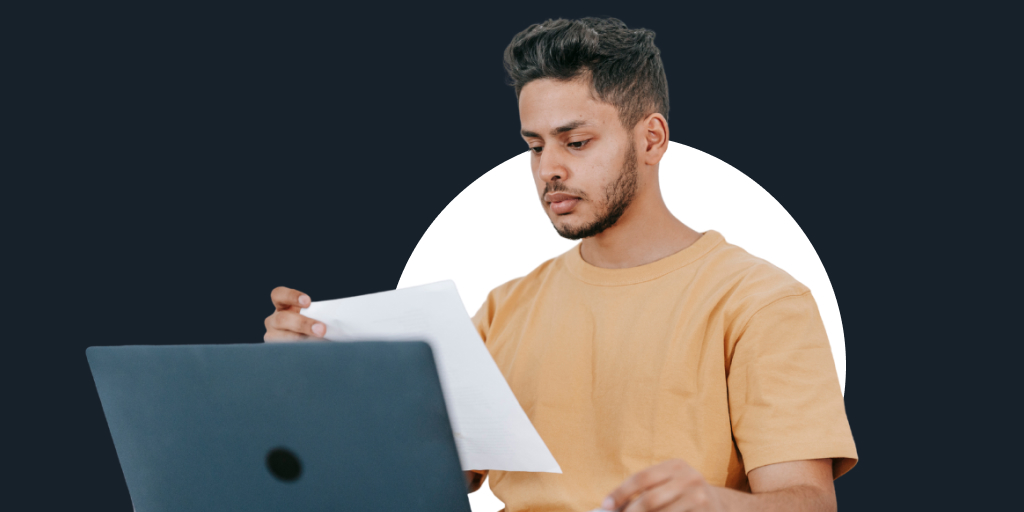 Man sitting at a desk with reading a paper while in front of a laptop