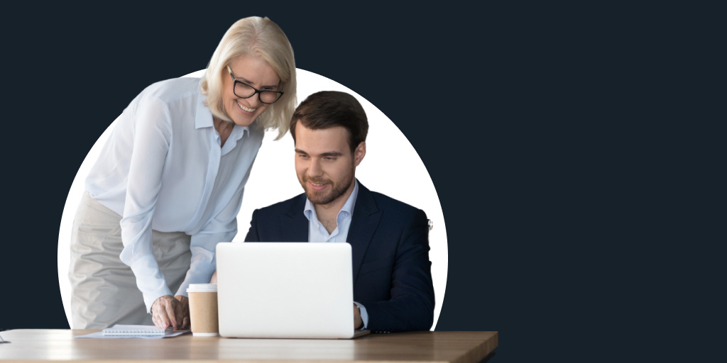 Two office professionals, a woman leaning over and helping a man working on a laptop