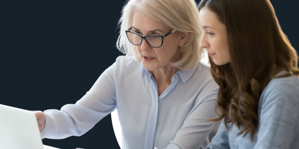 Two women, an older one mentoring a younger one in front of a computer