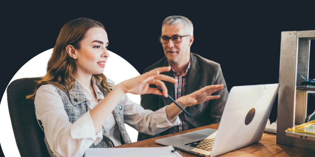 An older office worker helping out a younger employee as they discuss and work on a computer