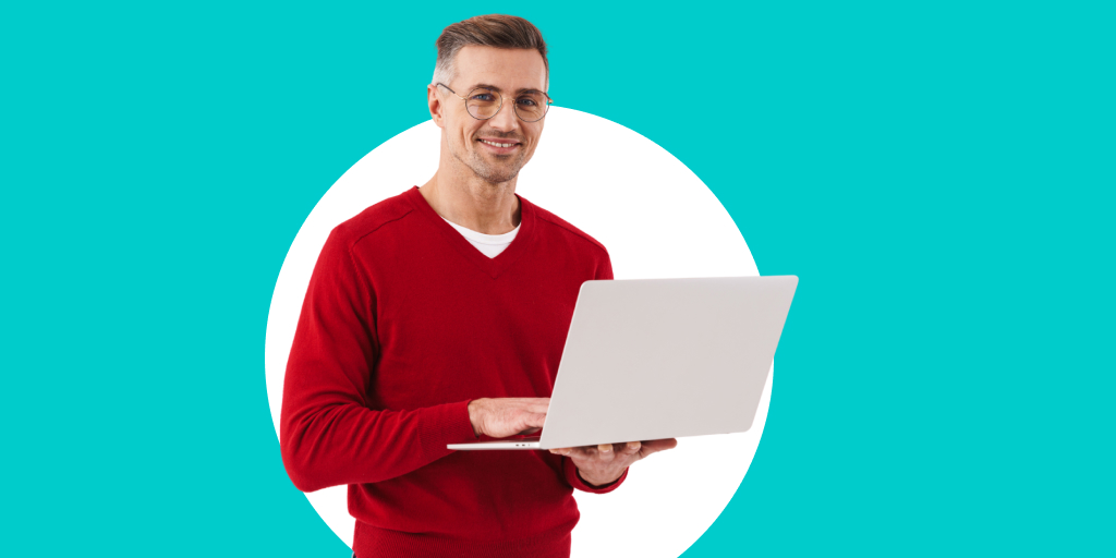 An older man with glasses, standing and smiling as he works on a laptop in his hands
