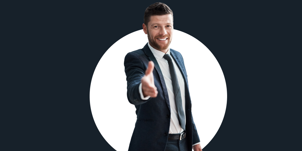 Man in a suit smiling with his hand out to shake, pointed at the camera