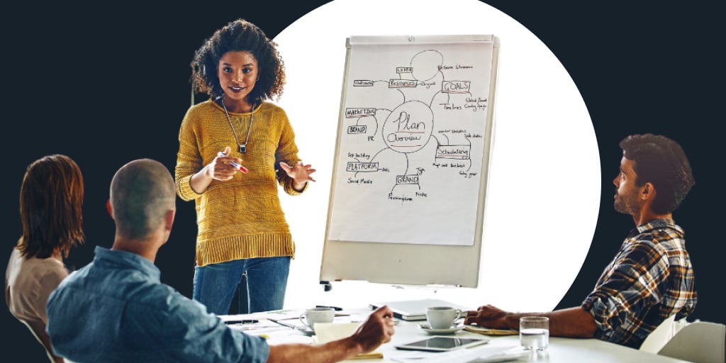 A group of people listening to a presenter who is referencing a whiteboard